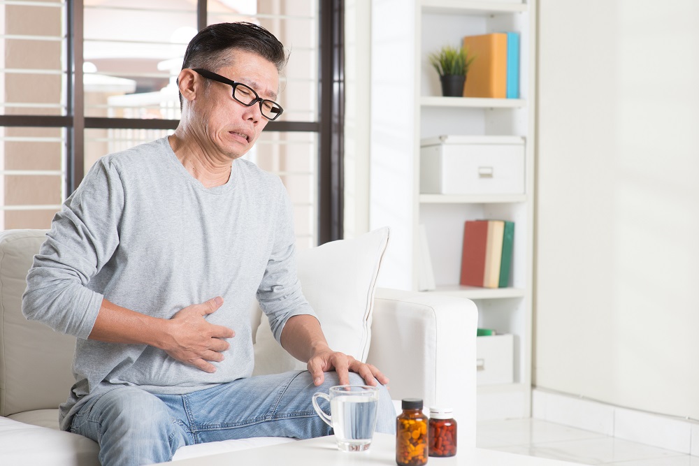 Mature 50s Asian man stomachache,  pressing on stomach with painful expression, sitting on sofa at home, medicines on table.